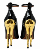 A pair of Alaia black patent leather high-heeled sandals shown from the back, featuring a distinctive metallic gold sculptural heel and ankle strap.