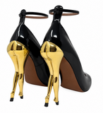 A pair of Alaia black patent leather high-heeled pumps with sculptural metallic gold stiletto heels and ankle straps, shown from the back on a white background.
