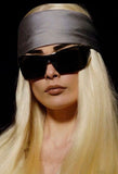 A close-up portrait of a model with long platinum blonde hair, wearing a wide gray satin headband and oversized black sunglasses against a solid black background. From Dior AW06 runway. 