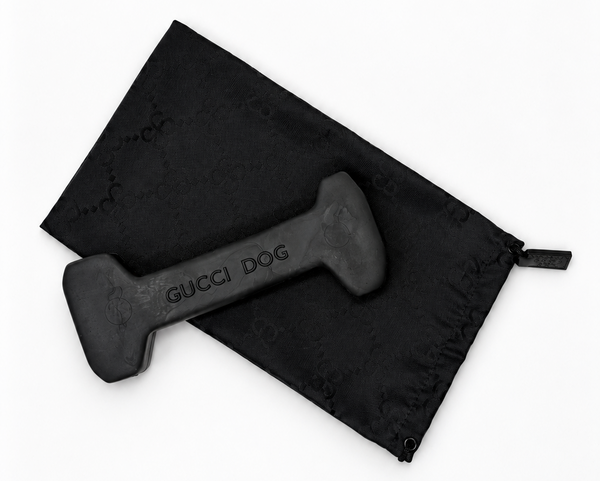 A black rubber dog toy shaped like a dumbbell with 'GUCCI DOG' printed on it, shown next to a black drawstring bag with a subtle Gucci monogram pattern on a white background.