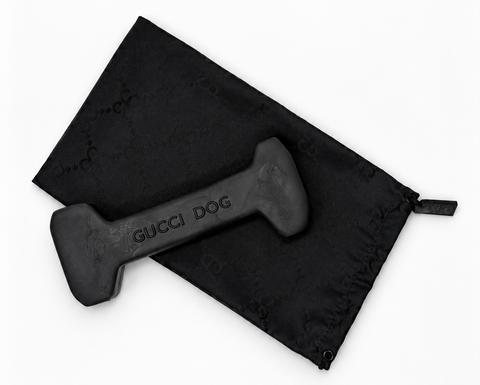 A black rubber dog toy shaped like a dumbbell with 'GUCCI DOG' printed on it, shown next to a black drawstring bag with a subtle Gucci monogram pattern on a white background.