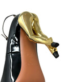 A close-up studio shot of a high-heeled shoe heel featuring a glossy black patent leather upper, a tan leather insole embossed with 'ALAIA PARIS', and a large sculptural metallic gold heel counter.