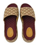 A pair of Gucci slide sandals in the classic GG canvas, featuring a beige and brown monogram pattern with burgundy straps and footbeds, shown from a top-down view on a white background.