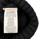 Close-up of a black ribbed knit or crocheted textile item with a sewn-on white product label reading shell/revètement, 100% wool/lana, and peacock feathers.