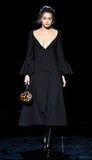 Gigi Hadid walks a fashion runway wearing a long black midi dress with a deep V-neckline, flared bell sleeves, and black knee-high boots, holding a leopard-print handbag against a dark stage background.