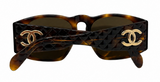 Chanel sunglasses with a dark brown and black tortoiseshell pattern, featuring a textured woven design on the front, gold-tone interlocking CC logos on the temples, and dark lenses.