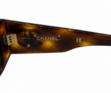 Close-up of the temple arm of Chanel sunglasses, showing the brand name 'CHANEL' and interlocking CC logo in a translucent brown tortoiseshell acetate on a white background.