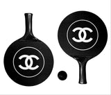 A pair of black paddle-shaped items with a carbon-fiber-like texture, each featuring a large white interlocking CC logo inside a circle, alongside a small black ball, on a plain white background. The logo is characteristic of Chanel.