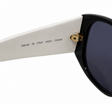 Close-up of a pair of sunglasses showing the white temple arm with black printed text, a gold hinge, and a black frame with dark lenses against a white background.