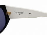 Close-up of a Chanel sunglasses arm showing the white acetate temple with black CHANEL branding, gold hinge detail, and dark tinted lenses against a white background.