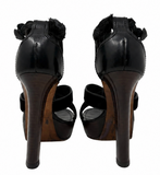 A pair of YSL black patent leather high-heeled sandals with ruffled straps and a brown wood-look heel, shown from the back on a white background.